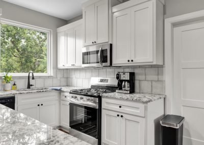 Modern kitchen with white cabinets, marble countertops, stainless steel appliances including a stove and microwave, a coffeemaker, a large window with trees outside, and a trash can by the door.