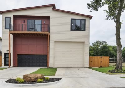 Modern two-story house with cream siding, black garage door, large beige garage door, small balcony, wooden fence, and a rock surrounded by plants in the front yard. Tree stands on the right side of the driveway.