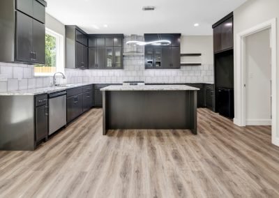 Modern kitchen with dark cabinets, granite countertops, a large island, stainless steel appliances, light gray tile backsplash, and wood-look flooring. Large window lets in natural light.