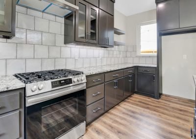 Modern kitchen with dark cabinets, stainless steel gas stove, marble countertops, light tiled backsplash, and wood flooring. Sunlight comes in through a window above the countertop.