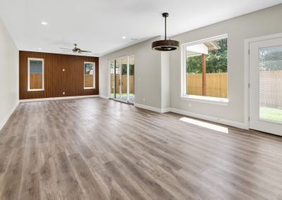 Spacious empty living room with wood flooring, large windows, sliding glass doors, ceiling fans, and natural light streaming in, overlooking a fenced backyard. Professional Real Estate Photography