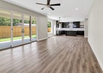 Open-concept living space with wood floors, large sliding glass doors, ceiling fan, and a modern kitchen featuring white tile backsplash, black cabinets, and stainless-steel appliances. The backyard is visible through glass doors—perfectly captured by a Tulsa Real Estate Photographer to showcase every detail for your real estate listing.