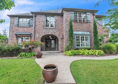 Two-story brick house with arched entryway, large windows, blue shutters, manicured lawn, shrubs, and two large empty planters along a curved concrete walkway under a bright blue sky—perfectly captured by a Tulsa Real Estate Photographer to showcase the property’s curb appeal.